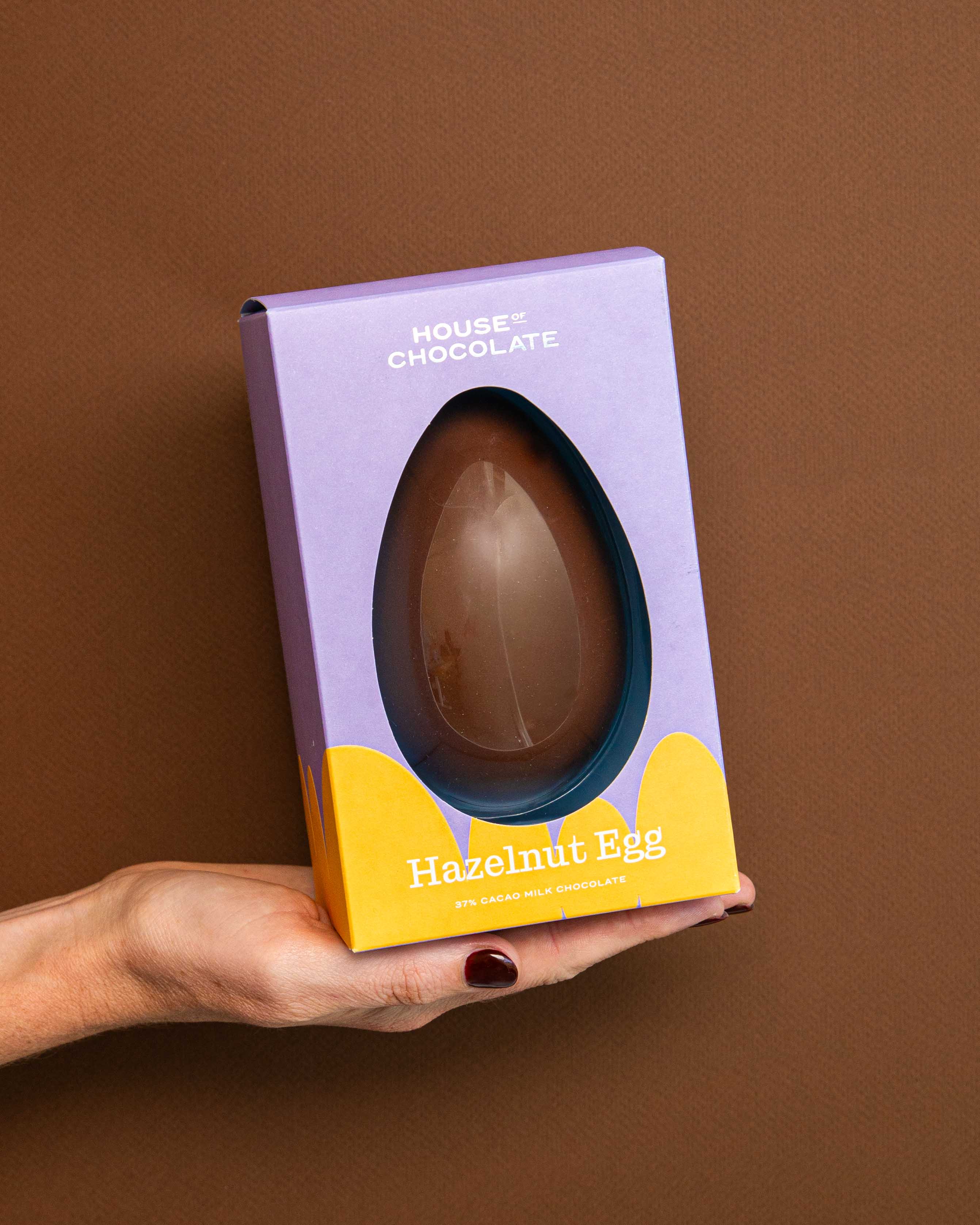 Hand holding a chocolate hazelnut egg in a purple and yellow box against a brown background