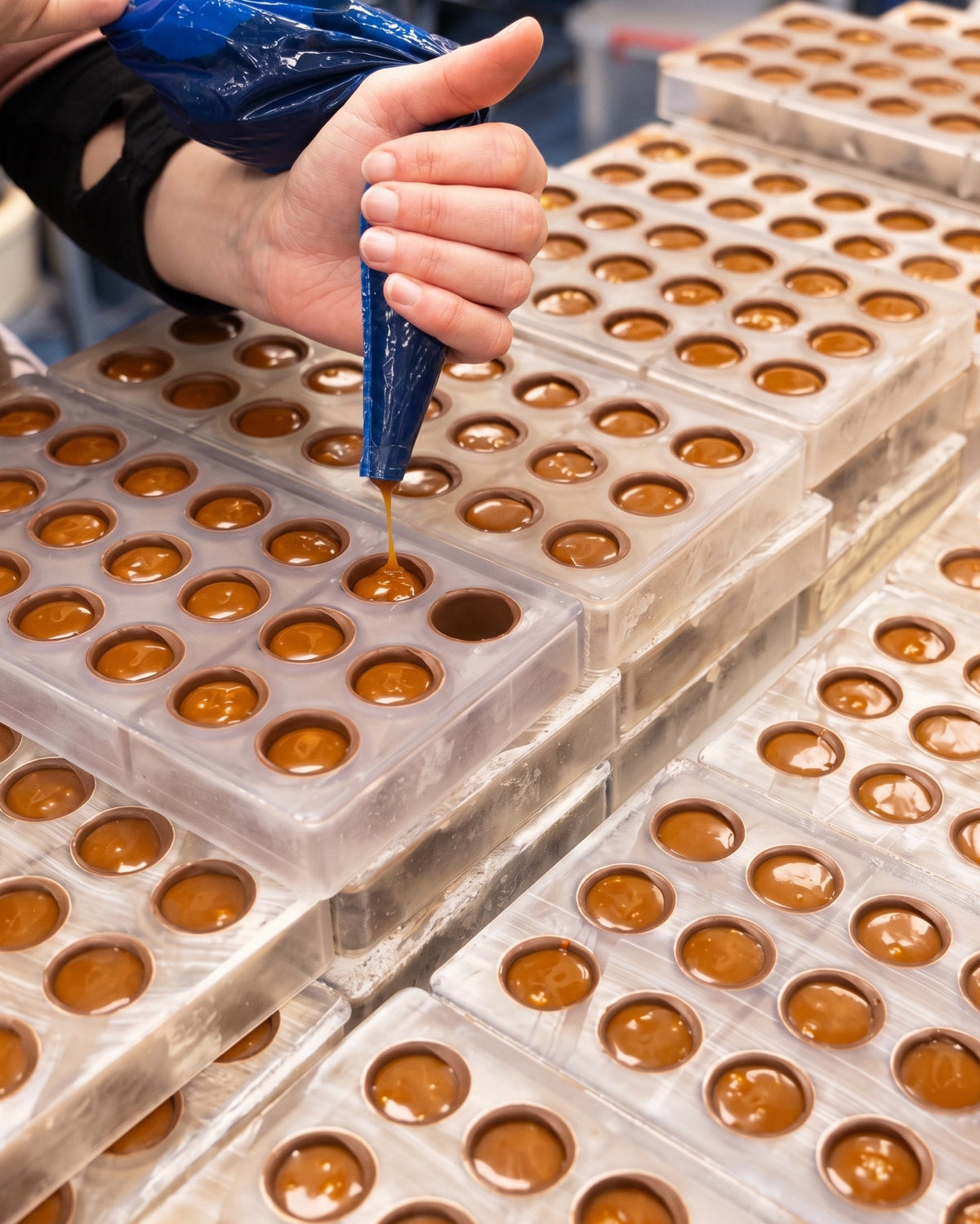 A close-up of a hand piping caramel into milk chocolate molds arranged in rows, with many similar filled molds visible in the background.